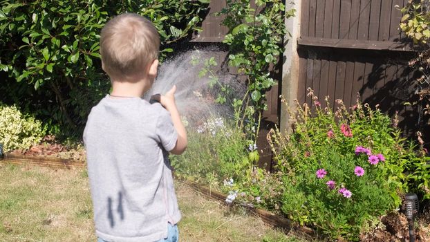 Funny little boy watering lawn plants in garden housing backyard. Adorable child playing with irrigation hose at hot sunny summer outdoors. Children help with housework. activity for kids. Childhood.