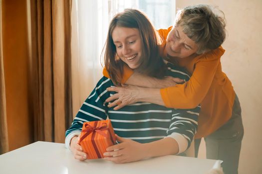 An elderly woman, a grandmother carefully hugs her granddaughter, gives her a surprise, makes a nice gift for the holidays, two women relatives have fun, a family happily spend time together.