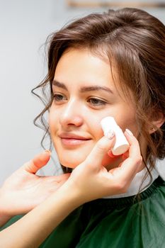 Makeup artist applying cream blush foundation tube on the cheek of the young caucasian woman in a beauty salon