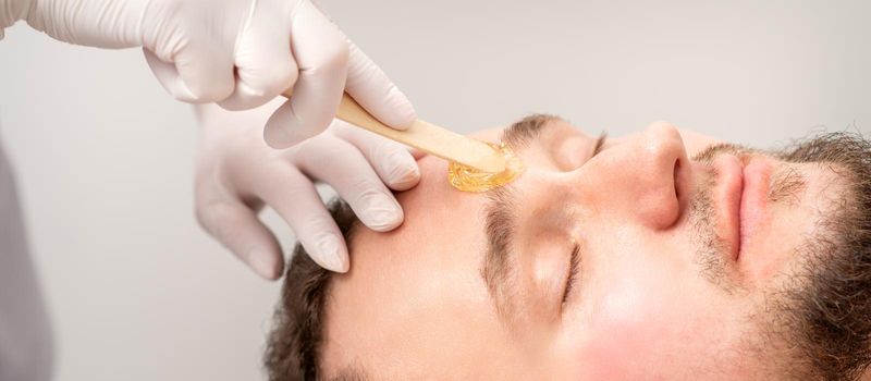 Beautician applying wax paste between eyebrows during the procedure of waxing in the beauty salon