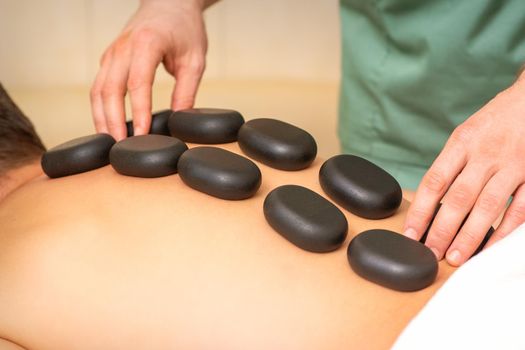Young caucasian man receiving back massage with black stones by masseur in spa salon. A man getting a spa treatment