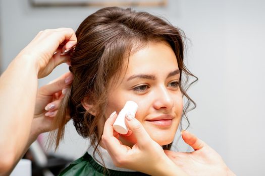 Makeup artist applying cream blush foundation tube on the cheek of the young caucasian woman in a beauty salon