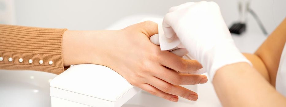 Hands of a manicurist in white protective gloves wipe female nails with a paper napkin in the salon