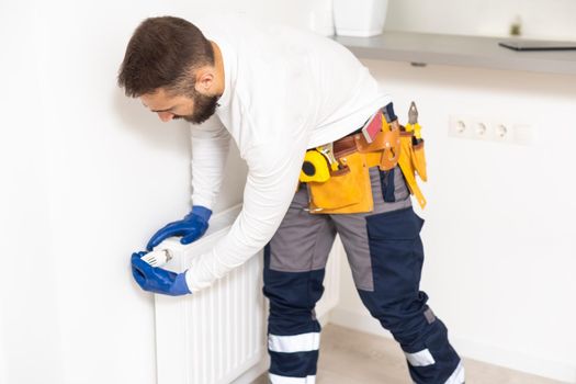 Man in work overalls using wrench while installing heating radiator in room. Young plumber installing heating system in apartment. Concept of radiator installation, plumbing works and home renovation