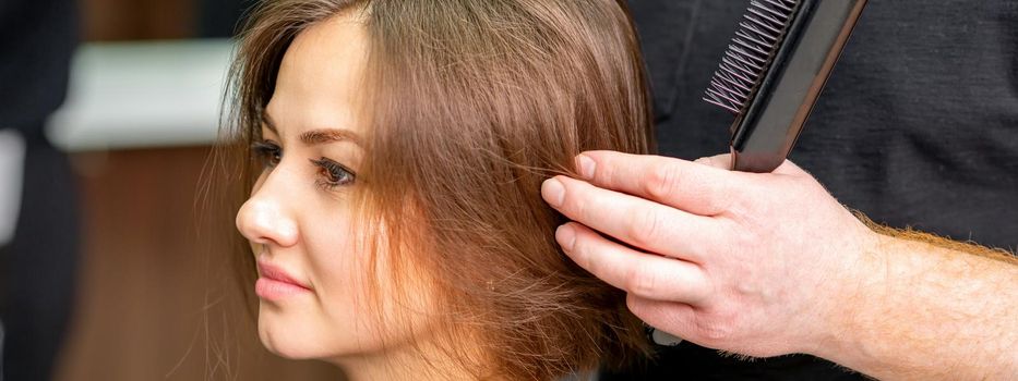 Male hairdresser works on the hairstyle of the young caucasian brunette woman at a hair salon