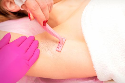 Beautician shaves the armpit of a young woman with a razor before the hair removal procedure