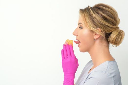 Young beautician licks liquid wax for depilation holding in gloved hands standing on white background. Cosmetologist tastes the sugar paste to taste