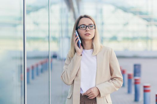 portrait of a serious woman wearing glasses blonde holding a mobile phone talking on a smartphone near the office