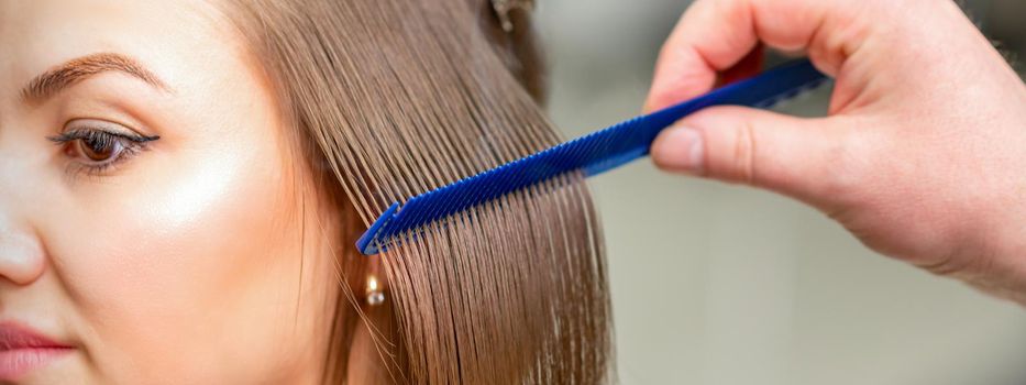 Hairdresser straightens female brown hair of medium length with a hair with an iron hair straightener and comb in a beauty salon