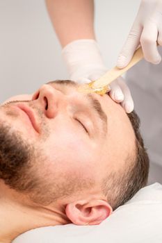 Beautician applying wax paste between eyebrows during the procedure of waxing in the beauty salon