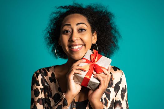 Excited african woman received gift box with bow. She is happy and flattered by attention. Girl smiling with present on blue background. Studio portrait. High quality photo