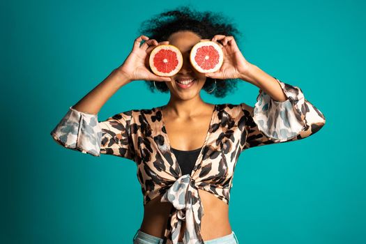 Pretty african american woman in leopard print top with two half of juicy grapefruit on blue studio background. Healthy eating, dieting, antioxidants concept. High quality photo