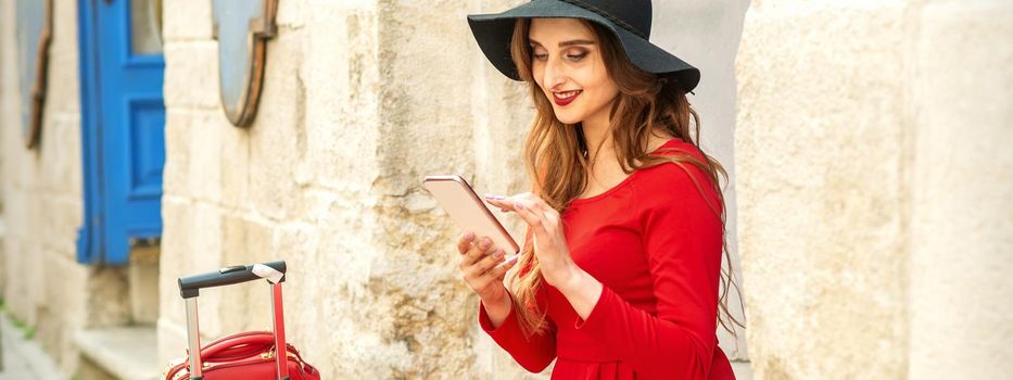 A young traveling white woman in the red jacket and black hat sitting with a smartphone on the city street