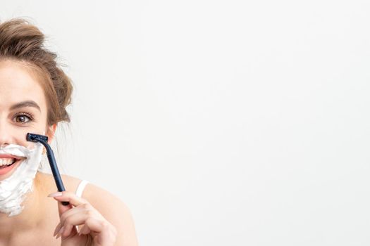 Beautiful young caucasian woman shaving her face by razor on white background. Pretty smiling woman with shaving foam and razor on her face