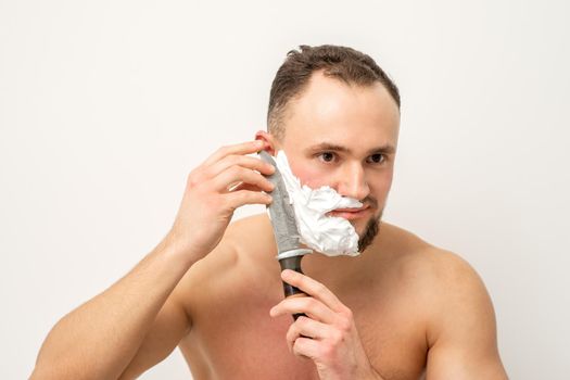 Young caucasian man shaving beard with a big knife on white background