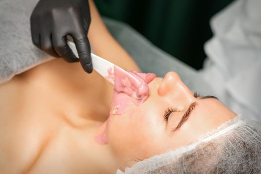 Beautician applying alginate peel-off powder facial mask with the spatula in a spa