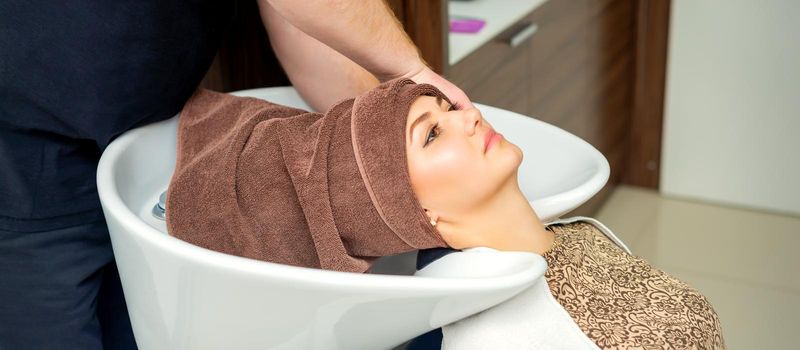 Gorgeous young relaxed woman getting her wet hair wrapped in a towel after washing by a professional hairdresser at the beauty salon