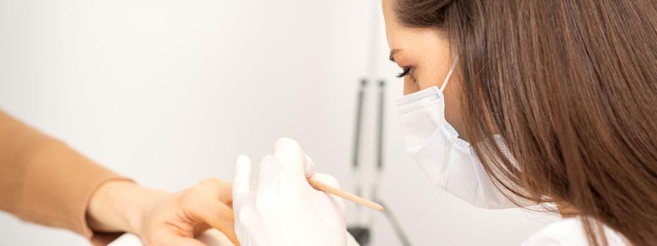 Manicure master in protective face mask and white gloves apply polish to female fingernails