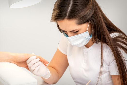 Manicurist wearing protective mask painting female nails with nail polish in manicure salon
