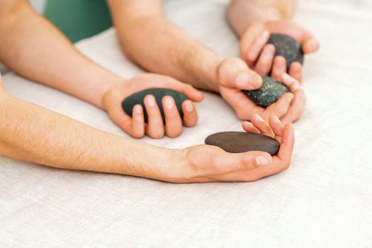 Four hands of male masseurs holding spa stones on white towel top view. Stone massage concept