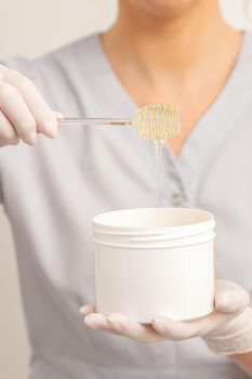 Liquid yellow sugar paste for depilation on a stick flows into the jar in hands of a beautician