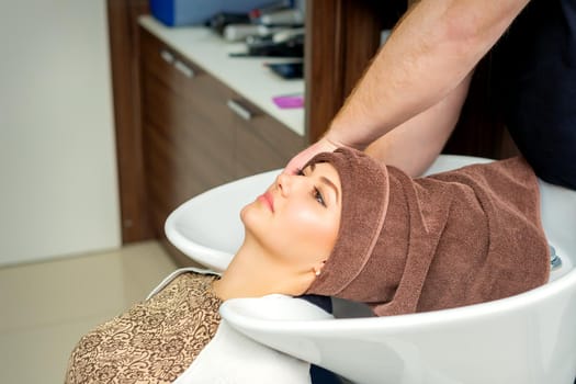 Gorgeous young relaxed woman getting her wet hair wrapped in a towel after washing by a professional hairdresser at the beauty salon