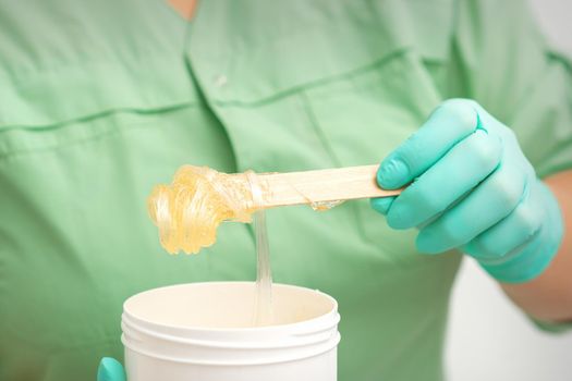 Liquid yellow sugar paste for depilation on a stick flows into the jar in hands of a beautician