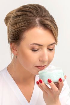 Portrait of young beautician sniffing moisturizing cream holding white jar on white background, copy space