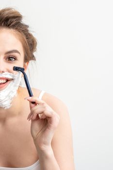 Beautiful young caucasian woman shaving her face by razor on white background. Pretty smiling woman with shaving foam and razor on her face