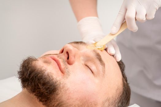 Beautician applying wax paste between eyebrows during the procedure of waxing in the beauty salon