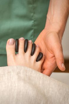 Feet massage with black pebbles between the female toes in hands of the masseur in the spa