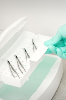 The beautician holds a tray of tweezers preparing them for disinfection with a special liquid and machine