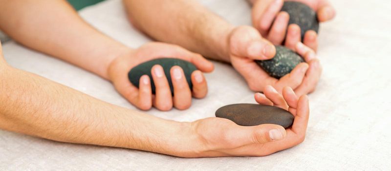 Four hands of male masseurs holding spa stones on white towel top view. Stone massage concept