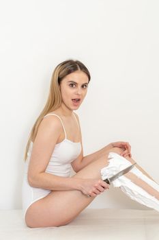 Beautiful caucasian woman shaving her legs with a knife with shaving foam on white background. Depilation and epilation concept