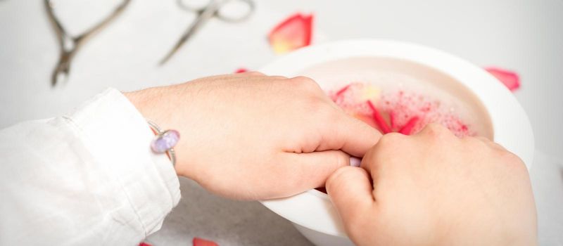 Hands in white bowl bathe nails with water and rose petals to soften the cuticle and dry skin