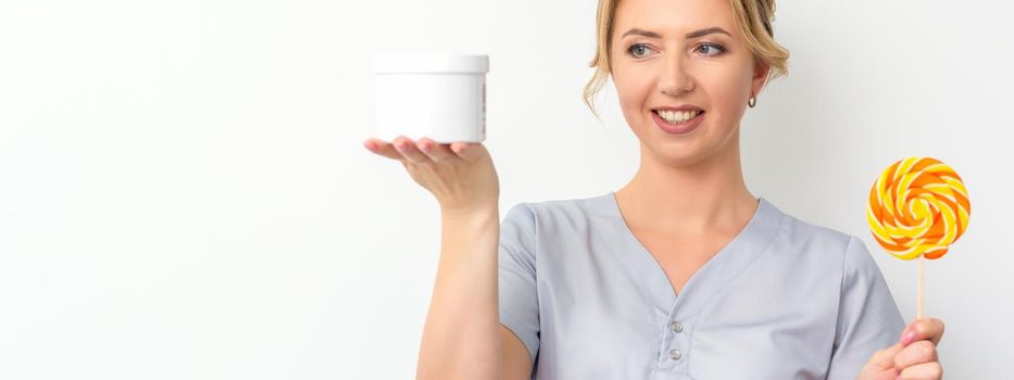 Beautician holding a jar of wax for waxing and candy on a stick smiling on a white background. Natural product for hair removal