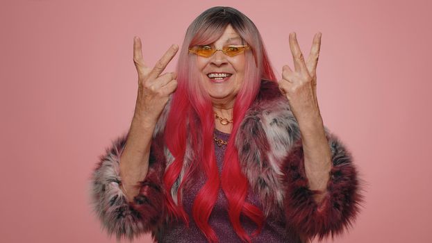 Happy joyful old woman in coat shouting, raising fists in gesture I did it, celebrating success, winning, birthday, lottery goal achievemen. Elderly grandmother indoors on pink studio wall background