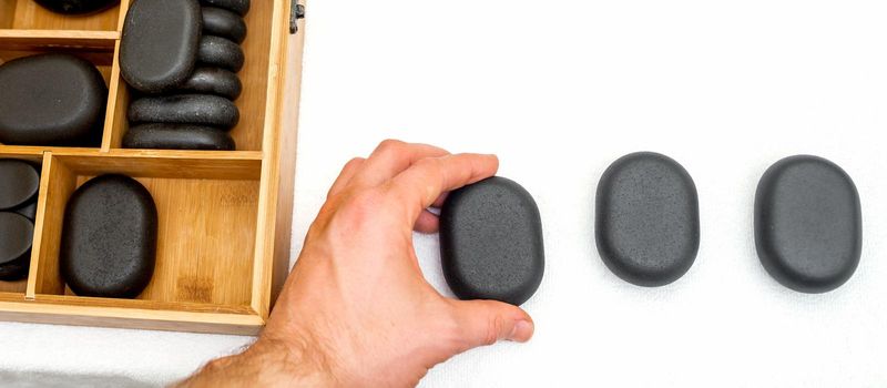 Hot stones for massage in a wooden box next to stone in the hand of masseur isolated on white background top view