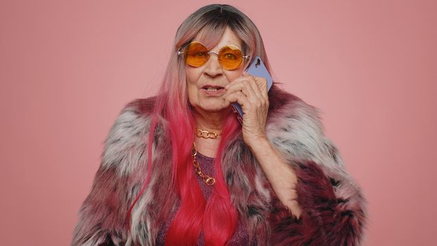 Senior elderly woman having pleasant mobile conversation using smartphone with friends, telling good news. Mature old grandmother making smartphone call isolated alone on pink studio wall background