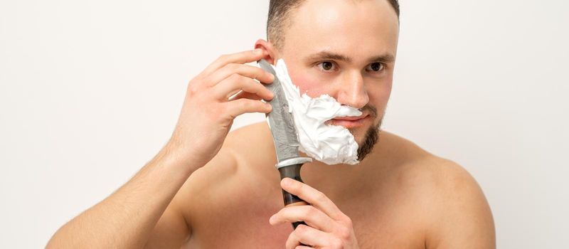 Young caucasian man shaving beard with a big knife on white background