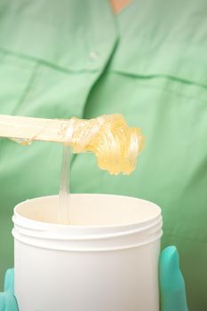 Liquid yellow sugar paste for depilation on a stick flows into the jar in hands of a beautician