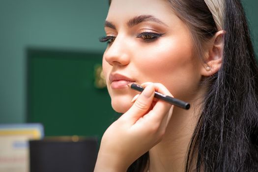 Makeup professional artist or cosmetologist is painting contour lips of a young woman with a pencil close up