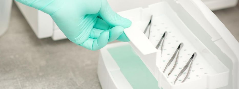 The beautician holds a tray of tweezers preparing them for disinfection with a special liquid and machine