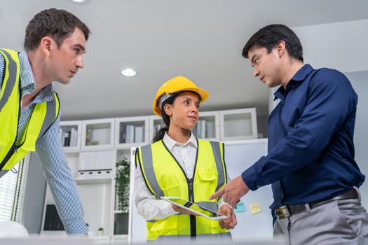 A team of competent engineers wearing safety equipment is working on blueprints with a tablet while also discussing with the investor in the office.