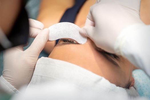 The beautician is gluing a patch under her eyes, before the procedure of eyelash extension. Artificial extended eyelashes