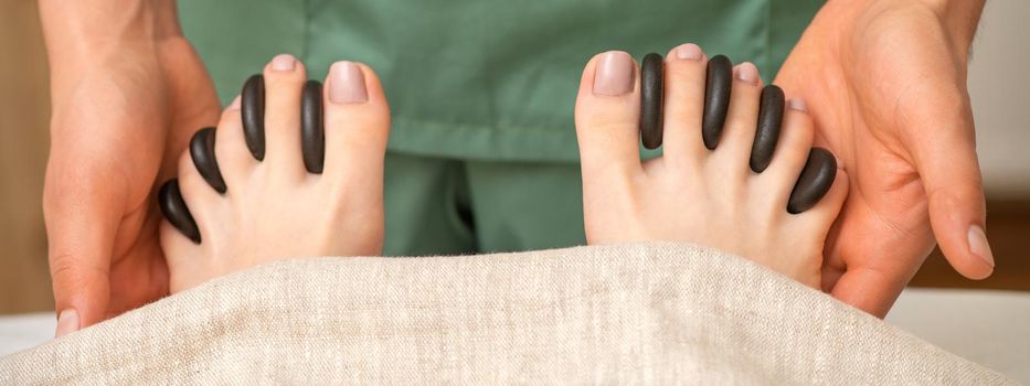 Feet massage with black pebbles between the female toes in hands of the masseur in the spa