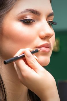 Makeup professional artist or cosmetologist is painting contour lips of a young woman with a pencil close up