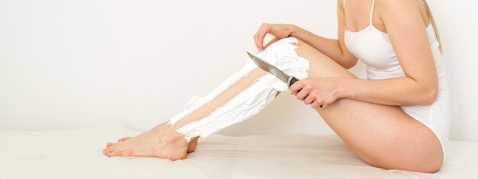 Beautiful caucasian woman shaving her legs with a knife with shaving foam on white background. Depilation and epilation concept