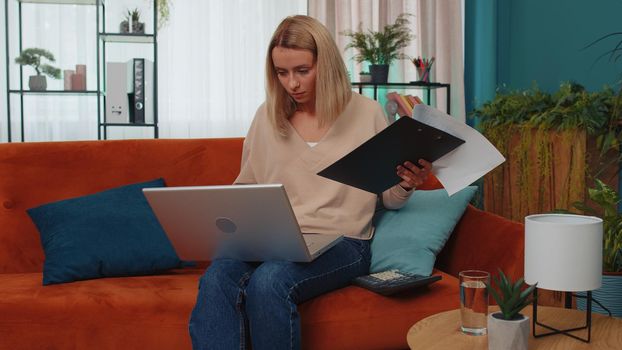 Girl accountant working with financial graph documents, calculate taxes for charts and graph reports of home income on couch. Young business woman working on laptop computer, planning expenses budget