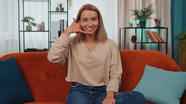 Call me, here is contact number. Adult caucasian girl looking at camera advertising doing phone gesture like says hey you call me back at home apartment. Young woman in living room sitting on sofa